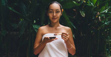 Makyaj fırçası ve doğal kremi olan genç Asyalı bir kadının portresi kameraya bakarken güzellik prosedürleri uyguluyor cilde ve vücuda önem veriyor canlılık hissi veriyor, çekici bir kadın pozu veriyor.