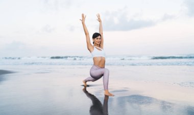 Sportif giyinen genç bir kadının portresi sabahları kameraya bakar kıyı şeridi sahilinde egzersiz yapar, çekici bir kızı incelerken pilates ve aerobik egzersizleri yapar.