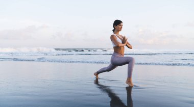 Haftasonu sahilde yoga yaparken günlük eşofman giyen sporcu bir kadın. Spor yapan bir kadın, deniz kenarında egzersiz yaparken vücut esnekliğini kontrol ediyor.