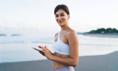 Dijital akıllı telefon teknolojisiyle kaygısız dişi Z kuşağının yarısı sahil şeridinde sabah gezintisinin keyfini çıkarıyor. Cep telefonu pozu veren büyüleyici blogcu portresi.
