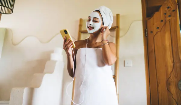 stock image Smiling Asian woman in headphones watching video on beauty blog while doing moisturize procedures in home, happy female in earphones using mobile app for reading article on cosmetology on website