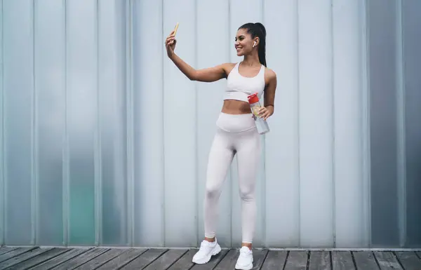 stock image Attractive healthy female in earphones photographing herself on smartphone camera, standing on copy space promo background. Young smiling woman blogger with fit figure posing for video on cellphone