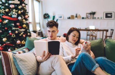 Asyalı kız evdeki kanepede akıllı telefon kullanırken beyaz adam kitap okuyor. Çok ırklı bir çift dinleniyor ve birlikte vakit geçiriyor. Noel ve yeni yıl zamanı. Ev eğlencesi ve eğlence.