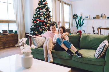 Evdeki kanepede Asyalı kız arkadaşıyla birlikte akıllı telefondan kitap izleyen neşeli beyaz bir adam. Çok ırklı bir çift birlikte vakit geçiriyor. Noel ve yeni yıl zamanı. Ev eğlencesi ve eğlence.