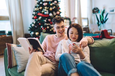Evdeki kanepede oturan Asyalı kız arkadaşına parmağını akıllı telefondan uzatan neşeli, beyaz bir adam. Çok ırklı bir çift birlikte vakit geçiriyor. Noel ve yeni yıl zamanı. Ev içi eğlence.