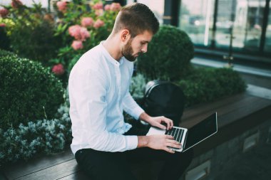 Beyaz gömlekli, siyah pantolonlu, tahta bankta oturan ve internette gezinirken dizüstü bilgisayarında yazan konsantre bir erkek.