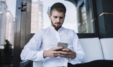 Resmi olarak odaklanmış sakallı erkek serbest çalışan koltukta oturup akıllı telefonla mesajlaşırken çağdaş kaldırım kafeteryasında çalışmayı düşünür.