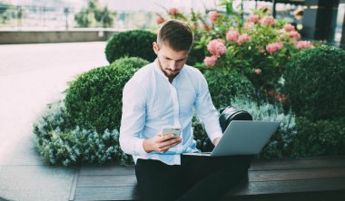 Beyaz tişörtlü, sakallı genç bir erkek, ahşap bankta otururken mesaj atıyor ve dizüstü bilgisayarında internette geziniyor.