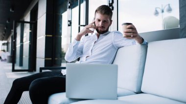 Ciddi bir iş adamı resmi kıyafetlerle kanepede dizüstü bilgisayar ve bir fincan kahveyle oturuyor ve çalışırken cep telefonuyla konuşuyor.