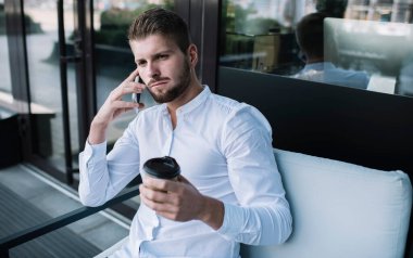 Resmi gömlekli düşünceli bir erkek işçi elinde bir fincan kahveyle caddede otururken ve akıllı telefonuyla konuşurken gözlerini kaçırıyor.