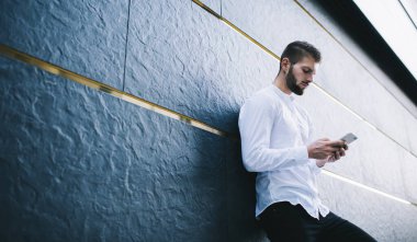 Beyaz tişörtlü, ciddi sakallı bir adamın modern binanın duvarına yaslanmış bir şekilde gündüz vakti akıllı telefon kullanmaya odaklanması.