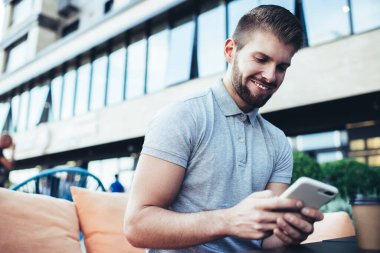Gündüz vakti verandada otururken ve arka planda bina inşa edilirken sıradan kıyafetlerle gülümseyen genç adam cep telefonuna mesaj atıyor.
