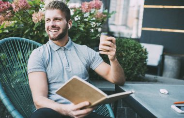 Keyifli bir adam, günlük kıyafetleriyle sokakta oturmuş, kahve içip kitap okuyor ve kablosuz kulaklıkla müzik dinliyor.