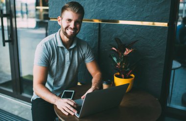 Pozitif erkek kameraya bakıyor ve netbook 'ta daktilo çekiyor. Terastaki açık kafede akıllı telefon ve saksıyla masada oturuyor.