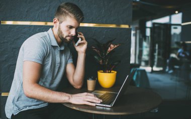 Düşünceli sakallı bir erkeğin, terasta açık bir kafede çalışırken akıllı telefon ve internet üzerinden konuşması.