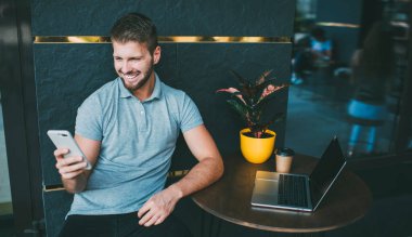 Gülümseyen, serbest çalışan, cep telefonuna göz atan, dizüstü bilgisayarla masaya oturan ve caddedeki modern kafenin terasında çalışan bir erkek.