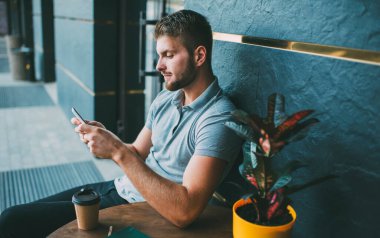 Düşünceli sakallı bir erkek, masada elinde kağıt bardak ve saksı bitkisiyle cep telefonu ekranına bakıyor.