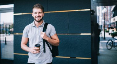 Mutlu erkek, dişlek gülüşü ve sırt çantasıyla modern binanın girişinde elinde kahve ile kameraya bakıyor.