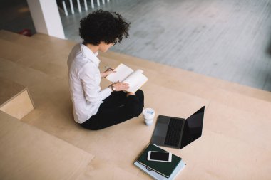 Bin yıllık bir kızın iş sırasında defterin sayfasını çevirmesinin en iyi görüntüsü. Modern başarılı kadın. Gözlüklü genç bir kadın ahşap bankta oturuyor. Elinde aletler, kitaplar ve kahve var. Çalışma odasında.