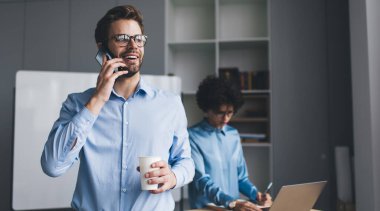 İş adamı elinde kahveyle akıllı telefondan konuşurken bayan meslektaşı dizüstü bilgisayarda yazı yazıyor ve masada not defteri yazıyor. Modern başarılı insanlar. Genç Kafkas milenyum insanları ofiste.
