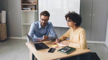 İş adamı dizüstü bilgisayarında, not defterine bir şeyler yazan bayan meslektaşına bir şey gösteriyor. İş birliği ve takım çalışması. Masadaki genç Kafkas milenyumu insanları. Modern başarılı insanlar