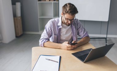Milenyum Kafkasyalı iş adamı iş sırasında akıllı telefondan mesaj atıyor ya da mesajlaşıyor. Modern başarılı adam kavramı. Ofisinde dizüstü bilgisayarını açmış, gözlüklü, utangaç genç bir adam.