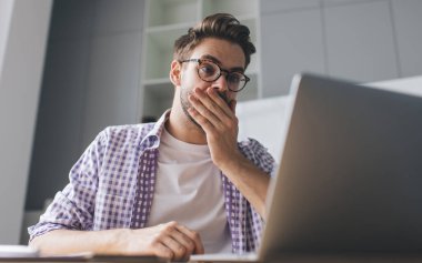 Bin yıllık beyaz iş adamının iş sırasında dizüstü bilgisayardan bir şey izlemesine şaşırdım. Modern başarılı adam kavramı. Ofiste otururken ağzını kapatan gözlüklü, şok olmuş genç adam.