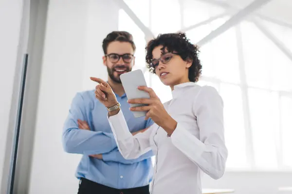 Stock image Businesswoman watching on smartphone and pointing with finger on interactive board to her male colleague. Concept of business cooperation and teamwork. Young caucasian millennial colleagues in office