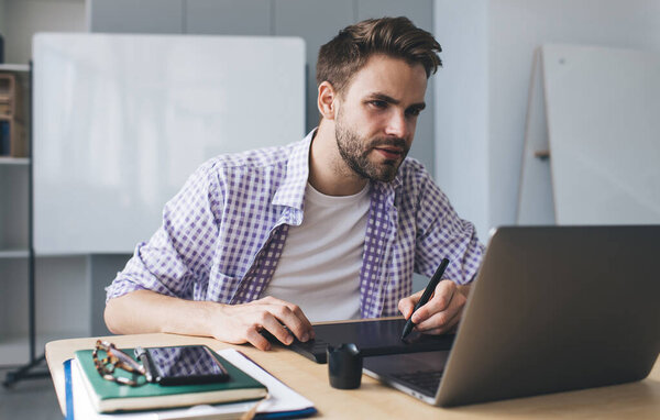 Millennial caucasian businessman watching on laptop and writing something in graphics tablet. Concept of modern successful man. Young concentrated guy sitting at desk in open space office