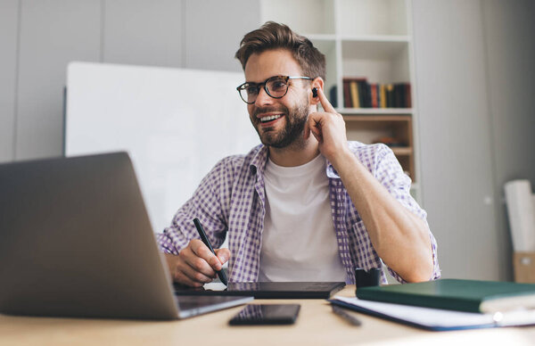 Millennial caucasian businessman working on laptop, writing in graphics tablet and correcting earphone in ear. Concept of modern successful man. Young smiling guy in glasses at desk in office