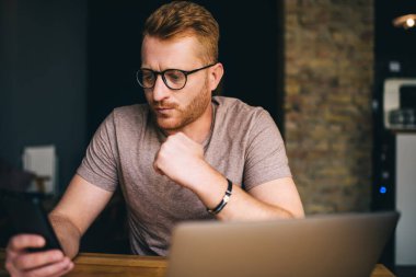 Beyaz saçlı, 30 yaşında, akıllı telefon uygulaması kullanan bir adam içeride dizüstü bilgisayar başında oturuyor. İş yerinde serbest çalışma sırasında cep telefonundan okunan şaşkın erkek mesajı