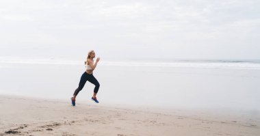 Tam uzunlukta, açık havada antrenman yaparken deniz kıyısında koşan beyaz tenli bir kadın. Genç bir kadın doğada spor yapıyor ve sağlıklı yaşam tarzının tadını çıkarıyor. Kaçak okyanus sahilinde hareket ediyor