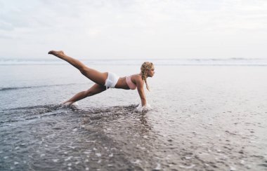 Uzunluk açısı geniş, spor vücutlu beyaz kadın açık havada antrenman yaparken deniz kıyısında esneme egzersizleri yapıyor. Genç bir kadın doğada spor yapıyor ve sağlıklı yaşam tarzının tadını çıkarıyor. Sahilde ısın.