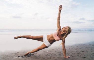 Uzunluk açısı geniş, spor vücutlu beyaz kadın açık havada antrenman yaparken deniz kıyısında esneme egzersizleri yapıyor. Genç bir kadın doğada spor yapıyor ve sağlıklı yaşam tarzının tadını çıkarıyor. Sahilde ısın.