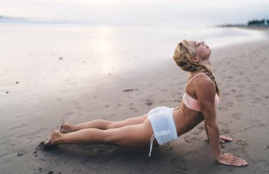 Uzunluk açısı geniş, spor vücutlu beyaz kadın açık havada antrenman yaparken deniz kıyısında esneme egzersizleri yapıyor. Genç bir kadın doğada spor yapıyor ve sağlıklı yaşam tarzının tadını çıkarıyor. Sahilde ısın.