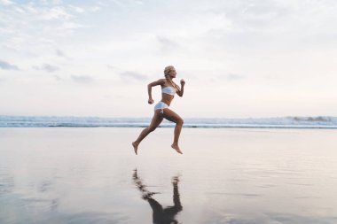 Tam uzunlukta, açık havada antrenman yaparken deniz kıyısında koşan beyaz tenli bir kadın. Genç bir kadın doğada spor yapıyor ve sağlıklı yaşam tarzının tadını çıkarıyor. Kaçak okyanus sahilinde hareket ediyor