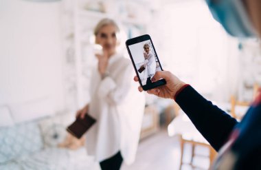 Bilinmeyen bir kadın tıbbi maske takıyor ve elinde akıllı telefonuyla gülümseyen Kafkasyalı bir kadının fotoğrafını çekiyor. Fotoğrafçılığın seçici odağı. Ev sanatları stüdyosunun iç görünümü bulanık