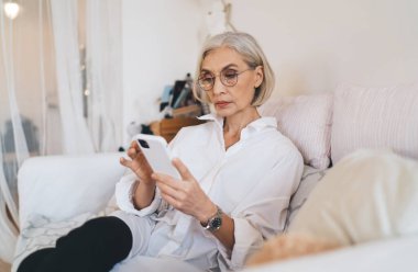 Konsantre olmuş, gözlüklü beyaz kadın koltukta oturuyor ve cep telefonundan bir şeyler izliyor. Ev istirahatı ve boş zaman kavramı. Modern geniş dairenin içi bulanık