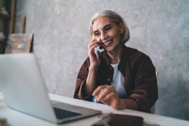 50 yaşında, beyaz, bayan girişimci, akıllı telefondan konuşan ve dizüstü bilgisayardan izleyen biri. Küçük işletmeler ve girişimcilik. Modern başarılı kadın ev sanatları stüdyosunda çalışıyor.