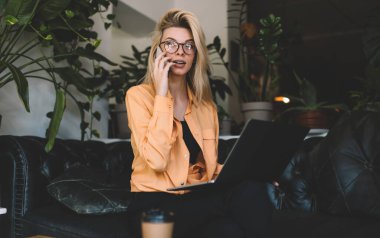 Netbook bilgisayarlı beyaz bir kadın cep telefonu konuşmaları sırasında içerik metnini tartışıyor, klasik gözlüklü bin yıllık hippi kız dizüstü bilgisayar programlama hakkında konuşmak için cep telefonuna sahip.