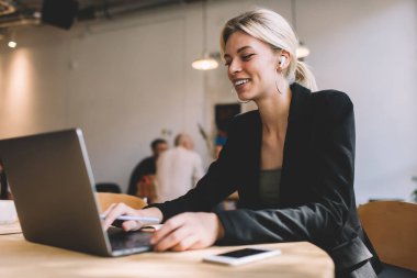 Kulaklıklı neşeli bir serbest yazar dijital netbook bilgisayarında profesyonel webinar videosu izliyor, Bluetooth kulaklıklı mutlu kadın grafik tasarımcısı ses dinliyor ve dizüstü bilgisayarda web sayfasını tarıyor.