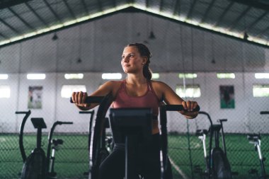 Eşofmanlı zayıf bir kadın spor aletleriyle spor yapıyor spor salonunda eğleniyor, spor yaparken spor yapan bir kadın sağlıklı yaşam ve zindeliğe ulaşmak için kardiyo eğitimi sırasında bisiklet sürüyor.