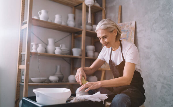 Smiling 50 years old european female sculptor making clay pot on pottery wheel. Concept of small business and entrepreneurship. Home hobby, entertainment and leisure. Woman working at art studio