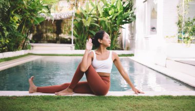 Balıkların Yarı Efendisi 'nde yoga yapan Asyalı kız dışarıda poz veriyor. Sağlıklı yaşam tarzı kavramı. Spor kıyafetleri giyen genç ve güzel bir bayan bahçede yeşil çimenler ve yüzme havuzu arasında oturuyor.