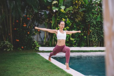 Savaşçı 'da yoga yapan beyaz bir kız. Açık havada poz veriyorum. Uyum ve akıl sağlığı kavramı. Spor giyimli genç bir kadın bahçedeki yeşil çimlerle yüzme havuzu arasında yalınayak duruyor.
