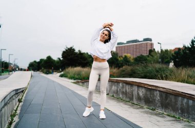 Spor kıyafetli neşeli bayan sporcu kameraya bakıp gülümserken taş kaldırımda durup yağmurlu bir günde şehir caddesinde ısınma egzersizleri yapıyor.