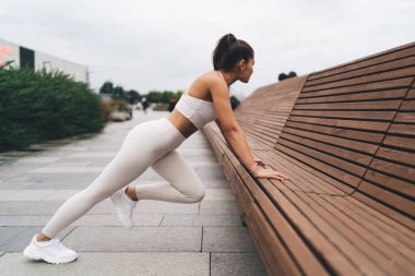 Beyaz spor kıyafetli genç bayan sporcu. Bulutlu bir günde şehir caddesinde antrenman yaparken tahta bankta yaslanıp bacaklarını esnetiyorlar.