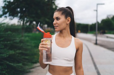 Uzun siyah saçlı, beyaz tişörtlü pozitif genç bayan yaz günü şehir sokaklarında egzersiz yaparken şişeden ferahlatıcı su içiyor.