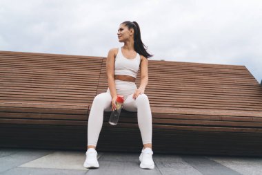 Bulutlu bir günde şehir parkında su dolu ahşap bankta dinlenirken gülümseyen ve başka yöne bakan, aktif kıyafetli genç bayan sporcu açısı düşük.