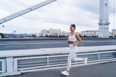 Spor kıyafetleri ve TWS kulaklıkları takan, gülen ve açık hava eğitimi sırasında köprüde koşarken şarkı dinleyen neşeli, genç İspanyol bayan sporcu.